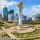 Aerial view of Astana's Bayterek Monument with surrounding plaza, modern skyscrapers, and landscaped gardens under blue sky